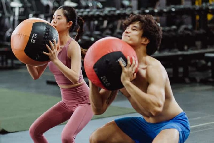 A man and woman exercising with medicine balls in a gym setting, focusing on strength and fitness.