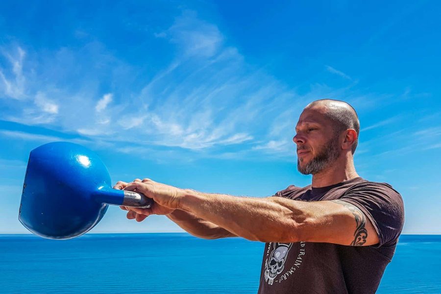 A fit, tattooed man lifting a kettlebell outdoors by the ocean under a clear blue sky.