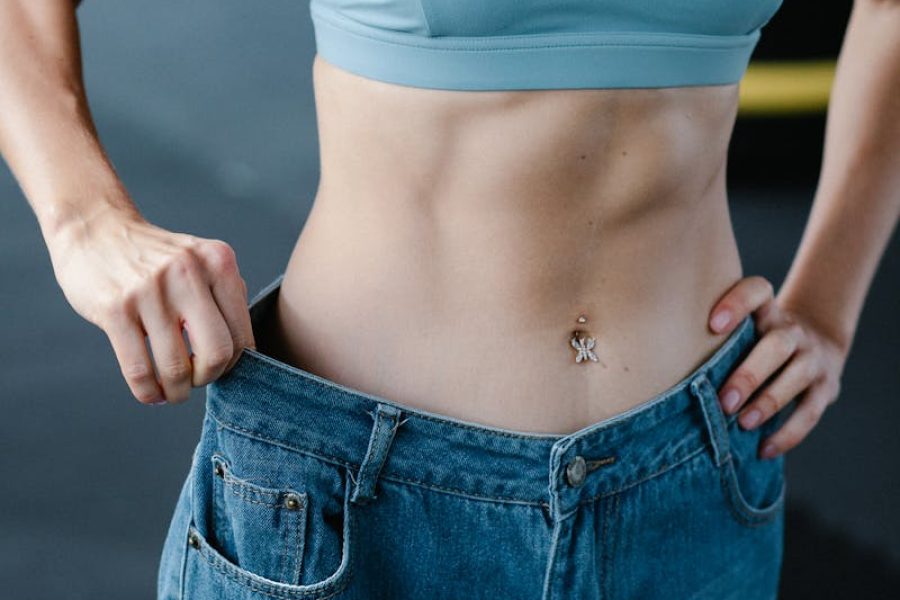 Fit woman shows weight loss in oversized jeans, displaying toned abs.